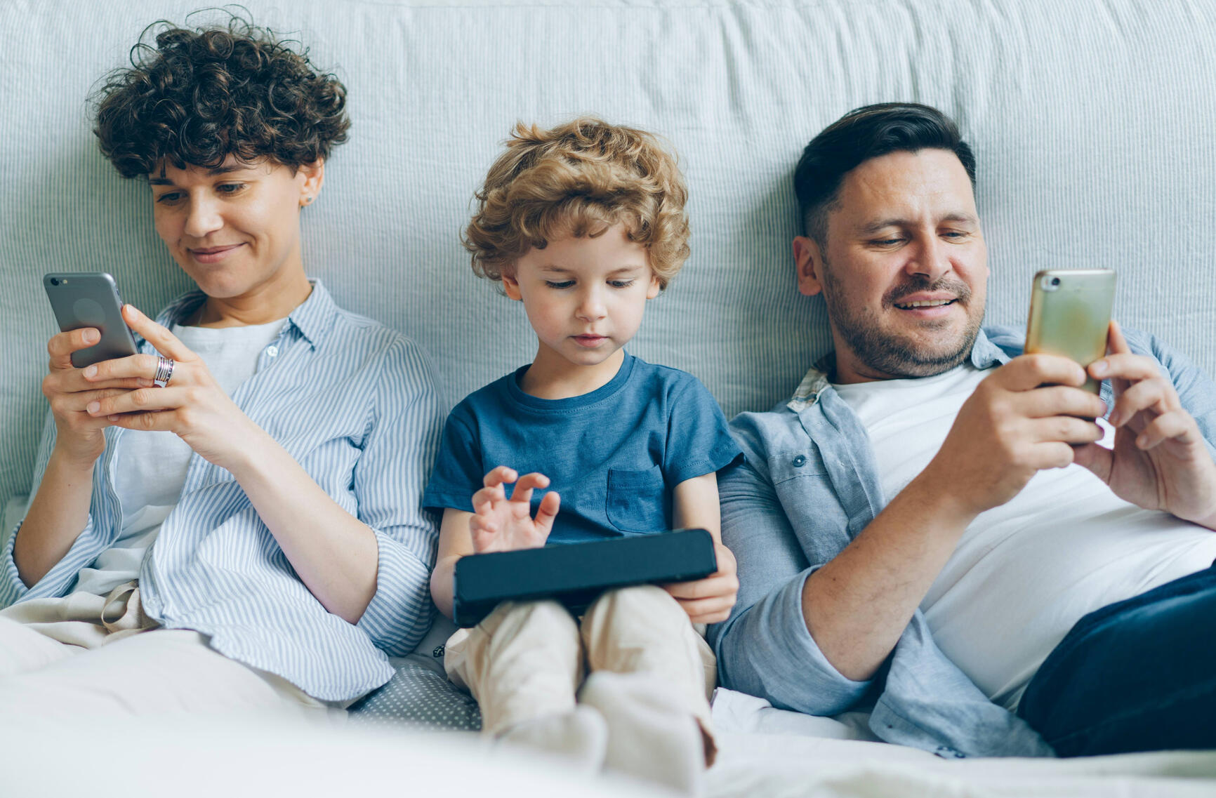 two adults and a child sit in bed, each using their own device